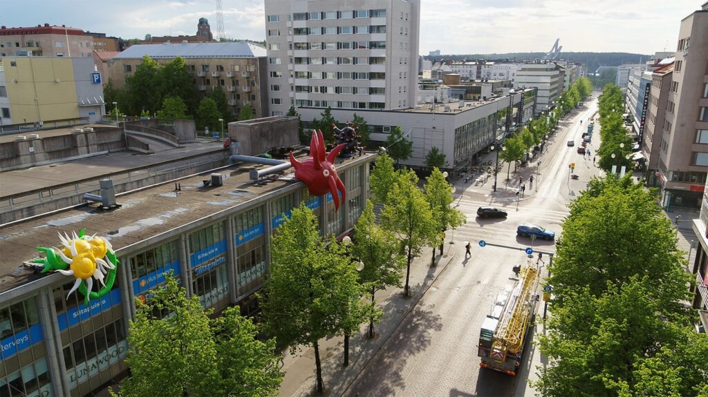 An aerial city view featuring three rooftop artworks on the left, in yellow, red, and black.