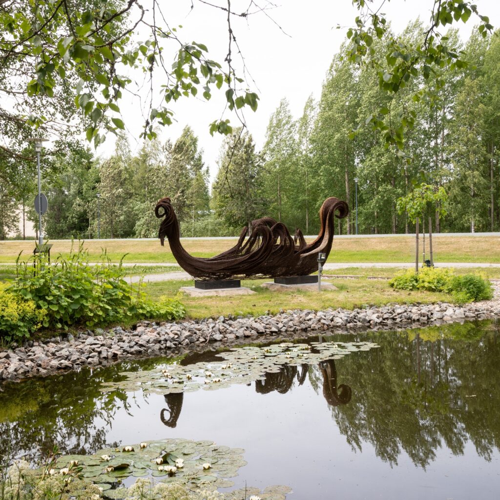 A large willow‑woven, boat‑shaped artwork by the water in a summer landscape.